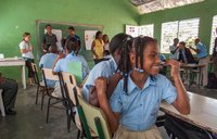 A girl dressed in a school uniform sits in a classroom, surrounded by other students and the teachers.