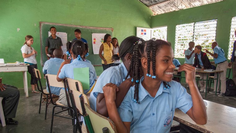 A girl dressed in a school uniform sits in a classroom, surrounded by other students and the teachers.