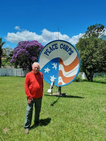 An older white male stands in front of a Peace Corps sign outside