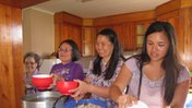Four Asian American women stand side by side in a kitchen. They are cooking together.