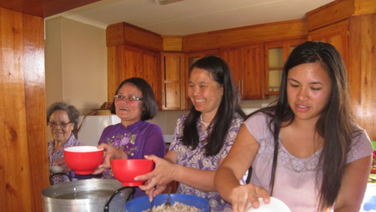 Four Asian American women stand side by side in a kitchen. They are cooking together.