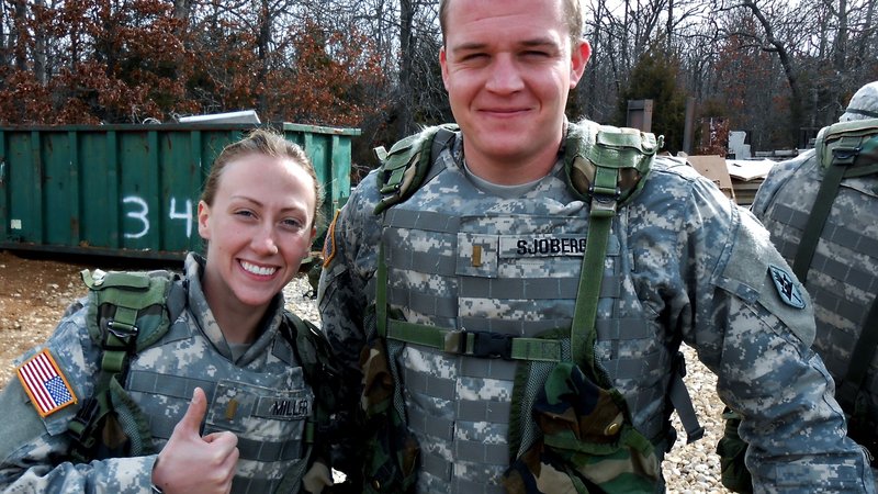 An American male in army fatigues and a female American in army fatigues stand next to each other smiling.