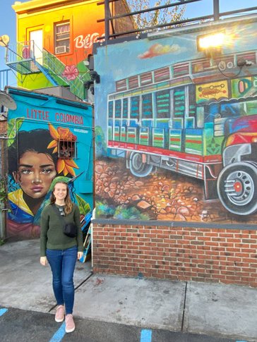A young woman stands in front of colorful street art that reads Little Colombia.