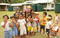 A Peace Corps Volunteer wearing a flower crown, stands with a group of smiling young children.
