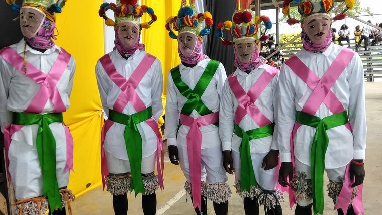 Five masked men wear pink and green sashes over an all-white outfit.
