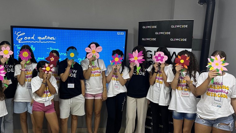 Twelve young students hold handmade paper flowers in front of their faces