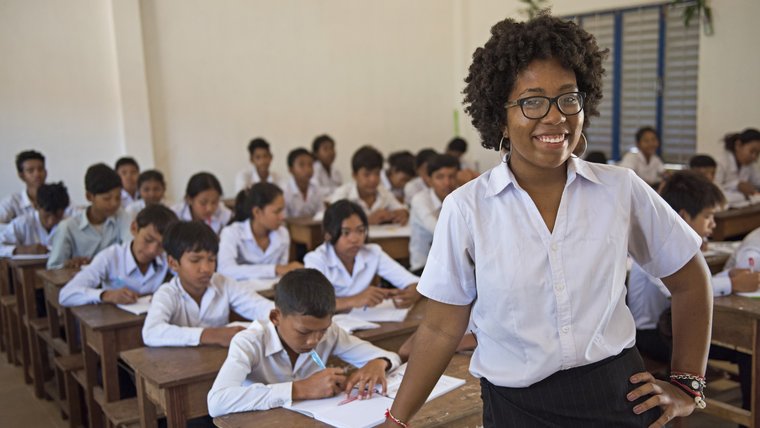 A Black woman leans on a school desk, smiling in front of her students, who are working on an assignment in the background
