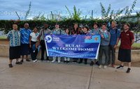 Twelve individuals stand behind a large banner saying "Bula! Welcome home"