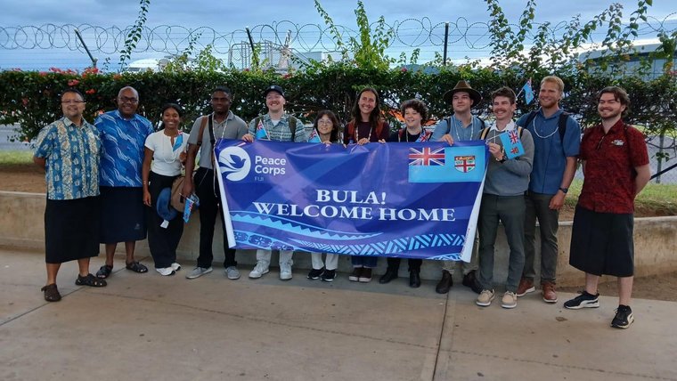 Twelve individuals stand behind a large banner saying "Bula! Welcome home"