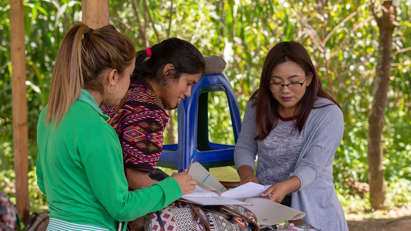 Hispanic female Peace Corps volunteer review paper documents with two female community members outside in Guatemala