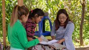 Hispanic female Peace Corps volunteer review paper documents with two female community members outside in Guatemala