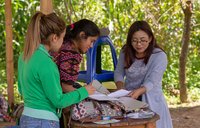 Hispanic female Peace Corps volunteer review paper documents with two female community members outside in Guatemala