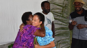 An Asian American Peace Corps Volunteer hugs a counterpart in Vanuatu. They are dressed in colorful clothing.