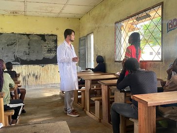 A white young man stands in a classroom talking to a female black young person, while other female and male young people are sitting