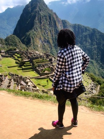 A Peace Corps Volunteer stands with her back to the camera. Machu Picchu, a bold mountain peak, is in the background.