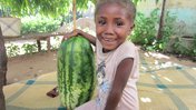 A young girl in Madagascar sits on a blanket next to a giant watermelon. She is smiling at the camera.
