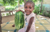 A young girl in Madagascar sits on a blanket next to a giant watermelon. She is smiling at the camera.