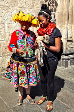 A Peace Corps Volunteer stands next to a woman in traditional Peruvian dress. The Volunteer is holding a baby sheep in her ar