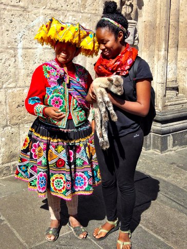 A Peace Corps Volunteer stands next to a woman in traditional Peruvian dress. The Volunteer is holding a baby sheep in her ar