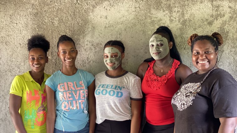 Five women stand together smiling. Two have mud masks on.