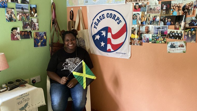 A young, black female smiles in her home in front of a Jamaican flag and a Peace Corps flag.