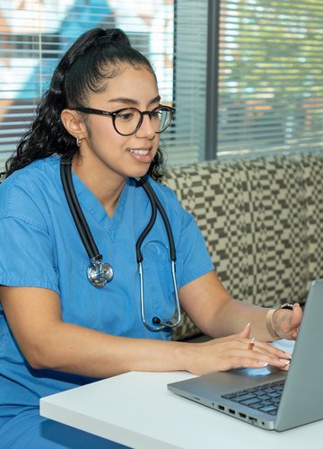 A female nurse in light blue scrubs sits in front of a laptop