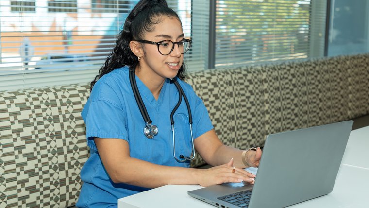 A female nurse in light blue scrubs sits in front of a laptop