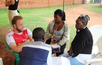 A male Volunteer sits with three Malawian campers under a tent outside.