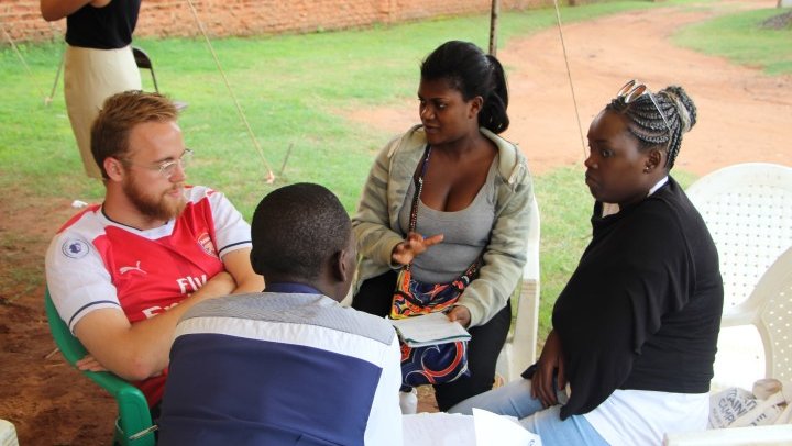 A male Volunteer sits with three Malawian campers under a tent outside.