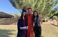 Three people stand together outside. The woman on the left and the man in the middle are wearing graduation gowns