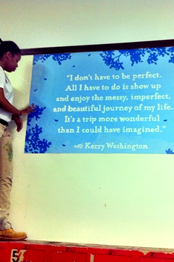 Brittany stands to the left of the frame on top of a cabinet. She is beside a freshly painted mural with a quote from Kerry W