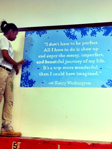 Brittany stands to the left of the frame on top of a cabinet. She is beside a freshly painted mural with a quote from Kerry W
