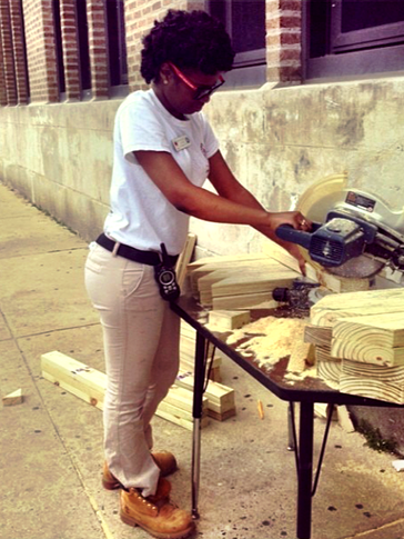 Brittany stands over a circle saw, cutting through pieces of wood on the sidewalk.