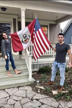 A man and a woman hold and American flag and a Peruvian flag outside of a house.