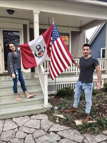 A man and a woman hold and American flag and a Peruvian flag outside of a house.