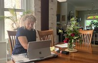 A woman sits at a dining room table working on her laptop