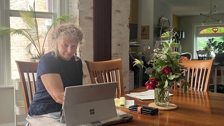 A woman sits at a dining room table working on her laptop