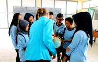 A female Peace Corps Volunteer stands in a circle of boys and girls in school uniforms, showing them something on a globe.