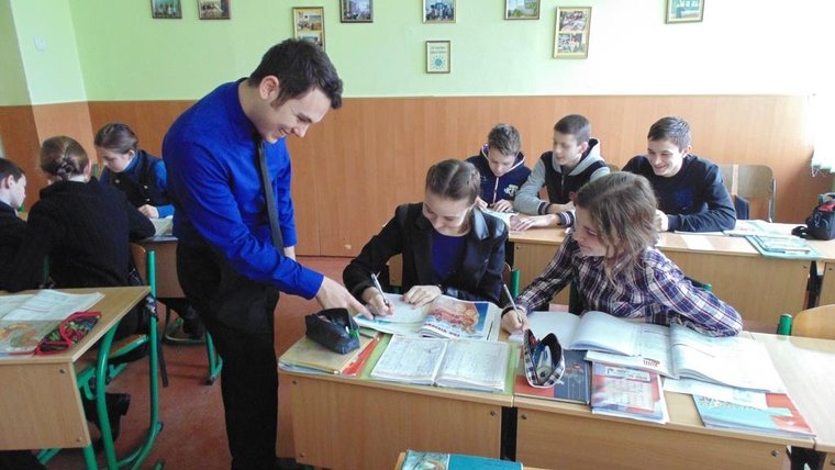 A Young white man leans over a desk to point something out to a younger student in Ukraine.