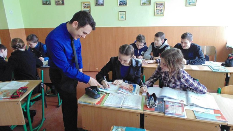 A Young white man leans over a desk to point something out to a younger student in Ukraine.