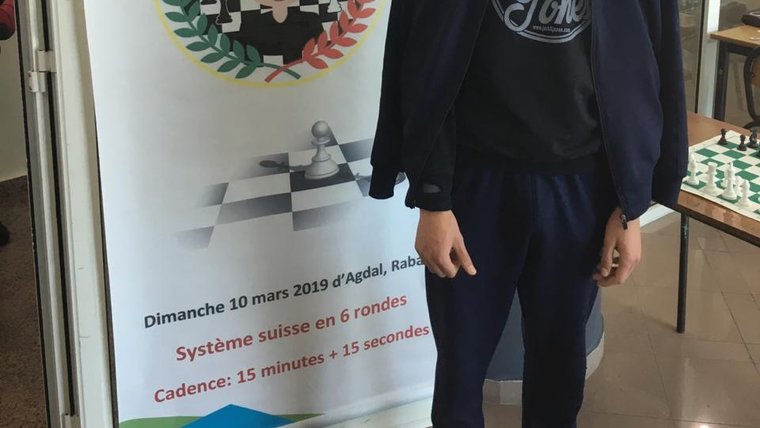 Ayman stands next to a large poster for a chess tournament
