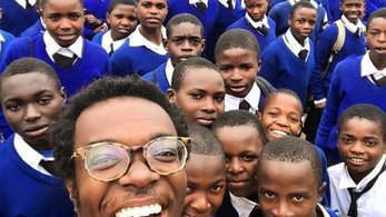 A young black man wearing round tortoise glasses takes a selfie with a group of Tanzanian students. The students are dressed 