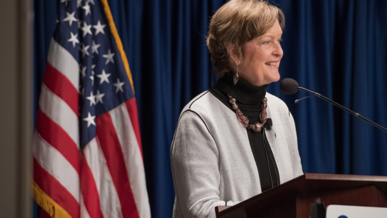 Jody Olsen stands at a podium that displays the Peace Corps logo