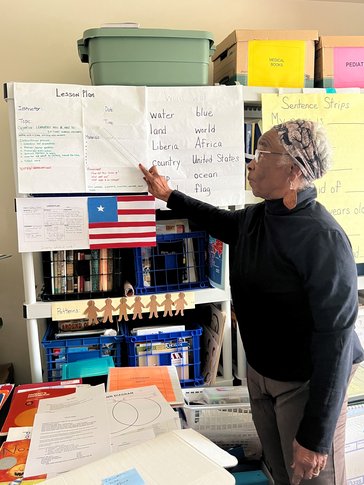 A woman points to a lesson plan on the wall with a ruler