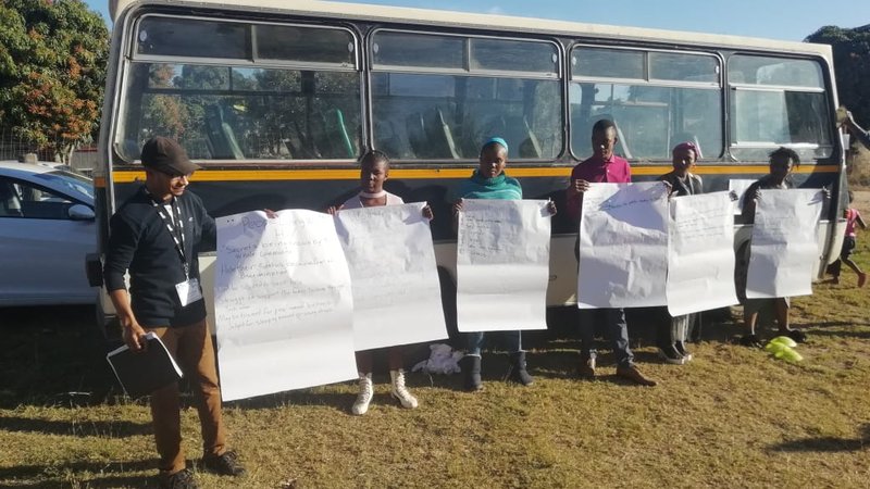 A Peace Corps volunteer stands with South African community members outside in front of a bus. There are flip chart papers ta