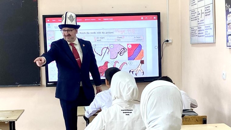 On “Kaplak Day” in Kyrgyzstan, David wears a special Kyrgyz hat and teaches a seventh grade class. A smartboard is in the background.