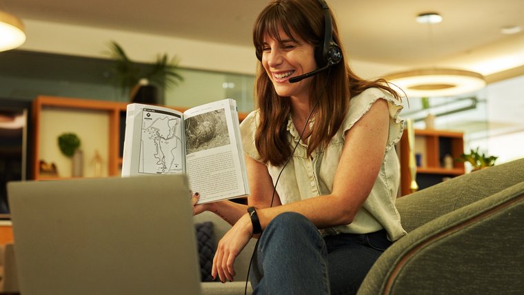 Woman smiles while reading a book in front of a laptop screen.