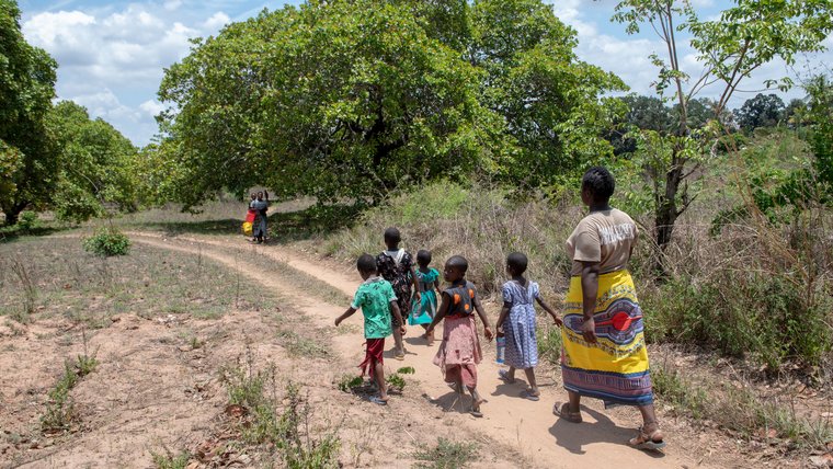 A Peace Corps Volunteer wearing a colorful, patterned skirt walks down a sandy path in Tanzania with a group of children.