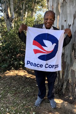 An older black man stands smiling with a Peace Corps flag in front of a tree.