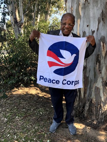 An older black man stands smiling with a Peace Corps flag in front of a tree.